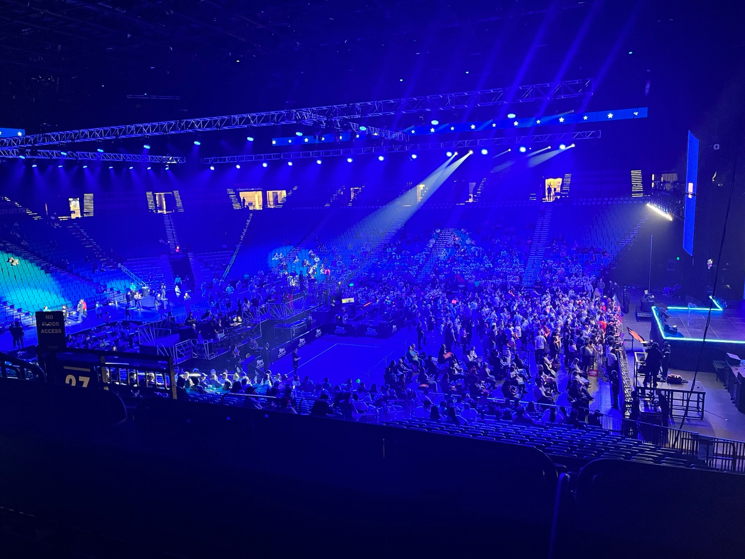 An arena audience and seating beneath blue show lighting and suspended production rigging