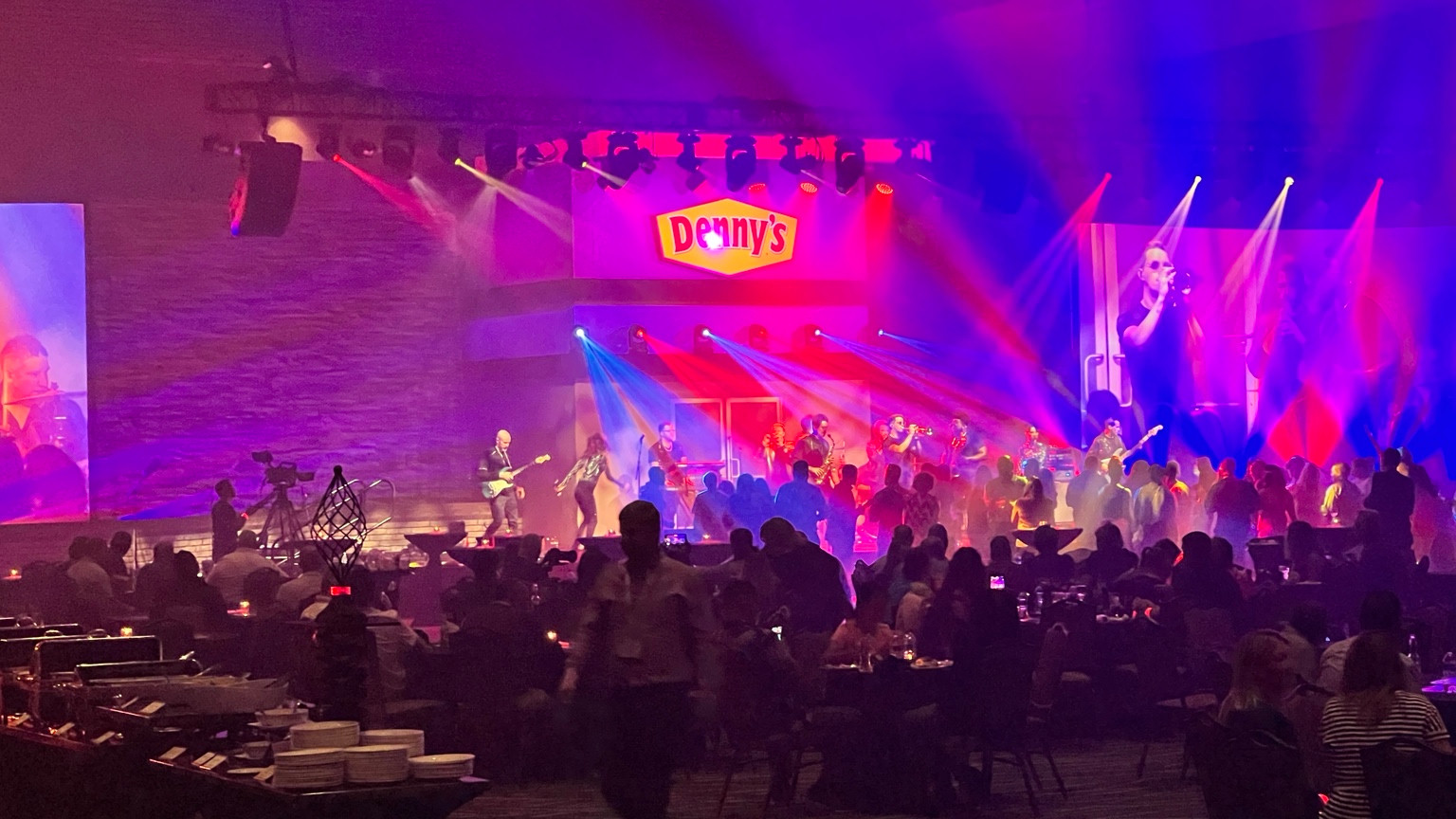 A Denny’s-branded stage with a live band, large video screens, vivid magenta and blue lighting, and an audience