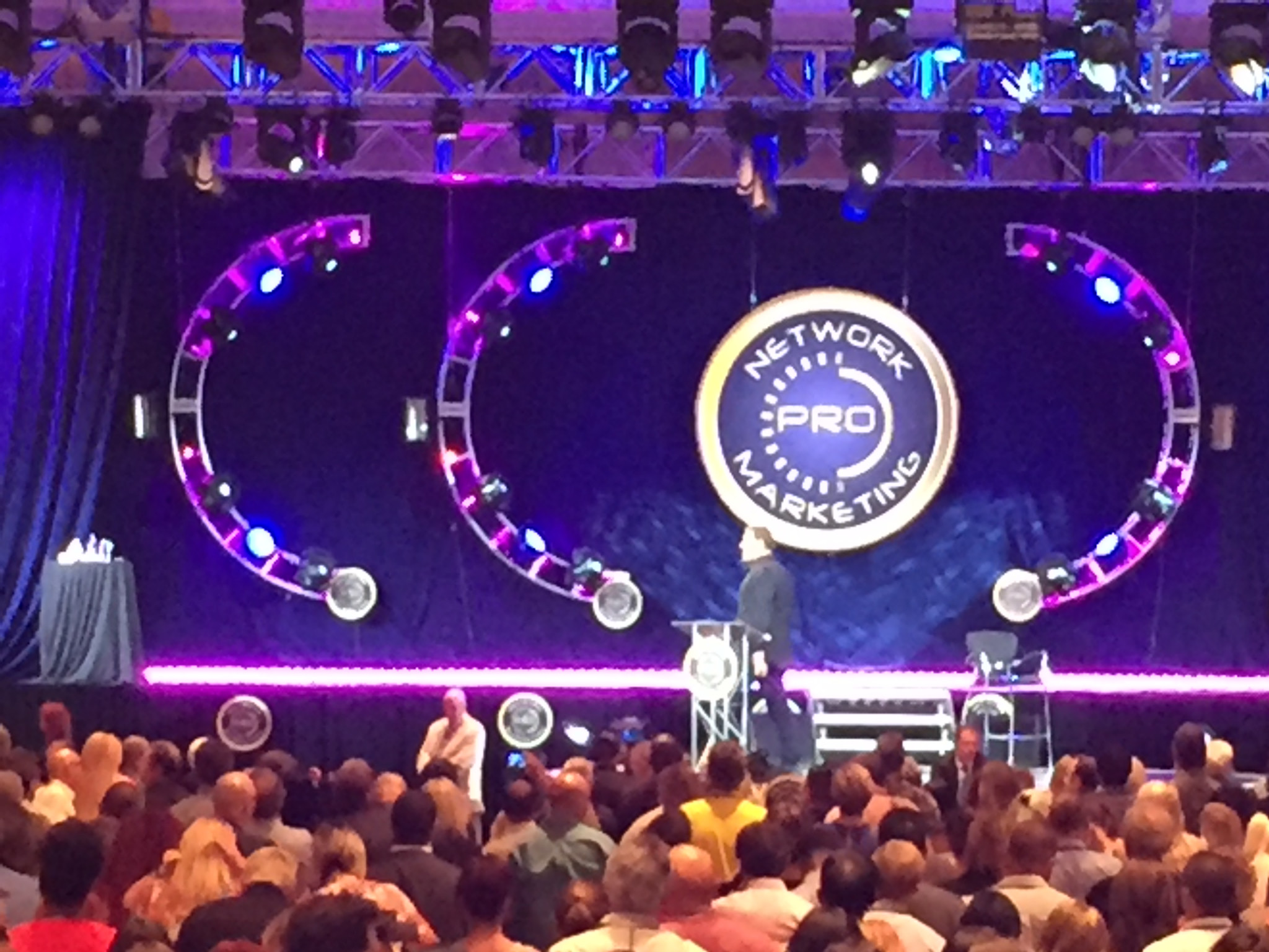 A Network Marketing Pro stage with three circular truss elements, purple lighting, and an audience